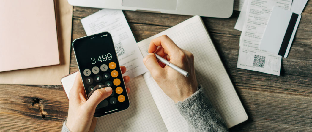 close-up of hands using the calculator app on a smartphone and writing in a notebook